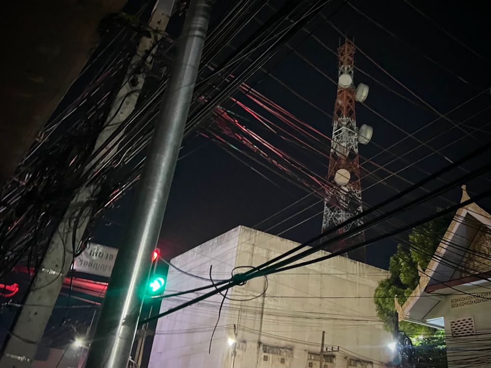 A telecoms tower in white and red with crisscrossed telegraph wires, partly lit red, in the foreground and a green traffic light at street level