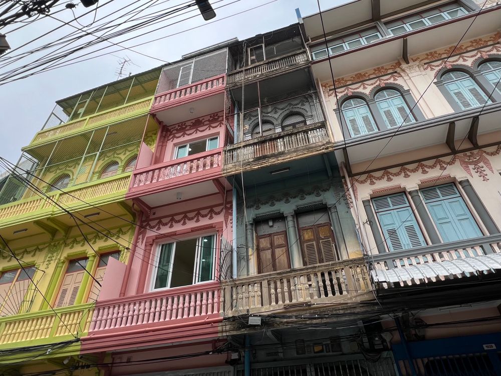 A row of four story city buildings, one green-yellow, one dark pink, one black/brown, and one pale pink, with balconies on each floor and shuttered windows