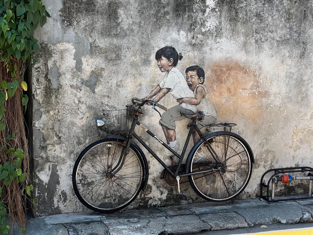 Old bicycle lent against a peeling street wall with a painting of two small, excited children looking like they’re riding the bicycle