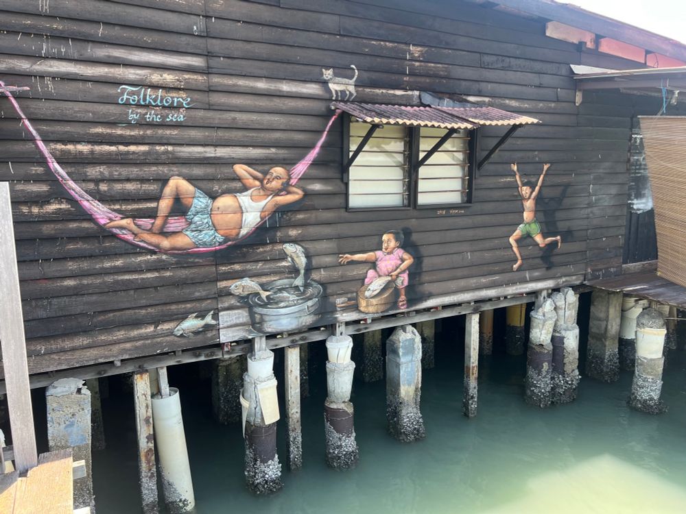 Street art depicting a boy leaping, a fat man sleeping in a hammock, a child sorting fish and a cat looking down, all painted on a wooden jetty building on stilts above the sea