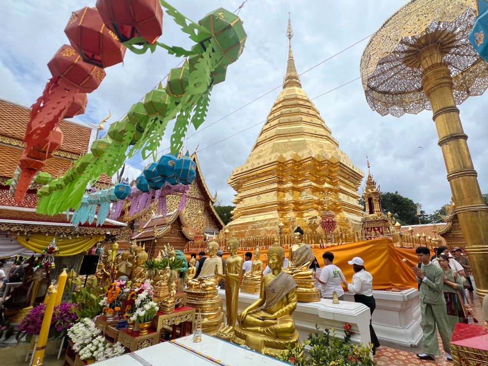 Buddhist temple with shining gold stupa, golden statues of the Buddha, and worshippers walking it around holding flowers. More flower offerings in the foreground along with red and green paper lanterns overhead