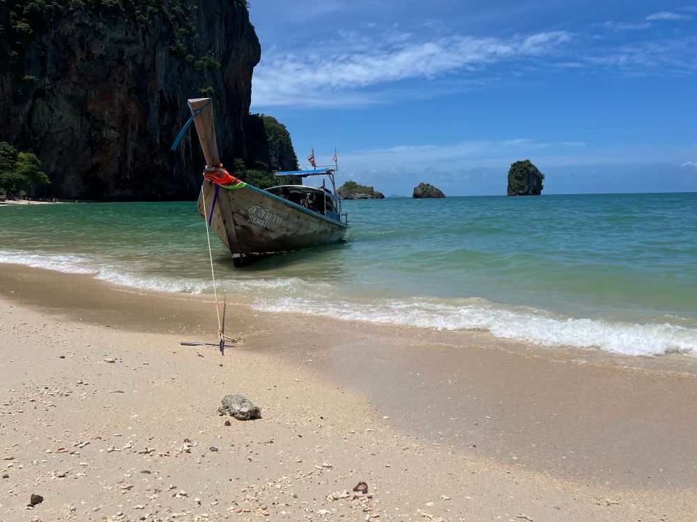 Wooden boat with long prow moored on a golden beach with green-blue sea in background, limestone cliffs, and small rocky islands