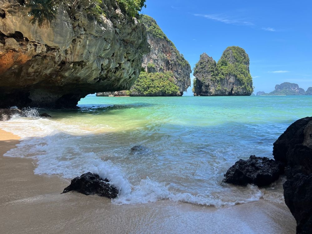 Wave breaking lightly onto sand between limestone rocks with green, rocky islands and turquoise sea in middle distance
