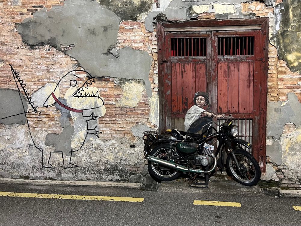 Street mural with a real motorbike leant against a pealing red doorway with a painted young boy in a helmet looking like he’s sitting on the bike