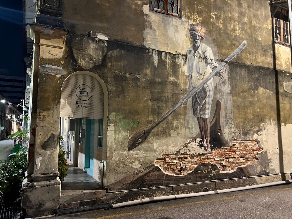 Realistic street mural of an old Indian man standing as he rows a wooden boat, painted on a old colonial building beside a covered wallet