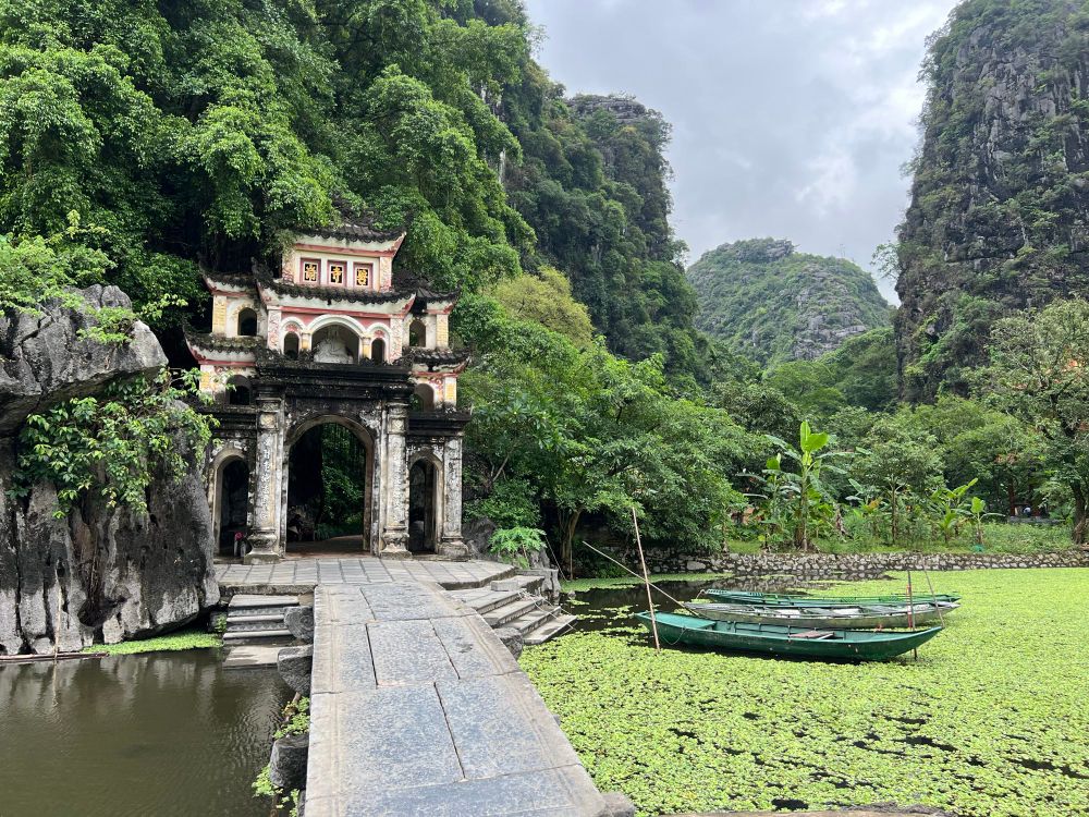 Buddhist gateway across a narrow stone bridge with water and boats beside the bridge and lush green hills in the background
