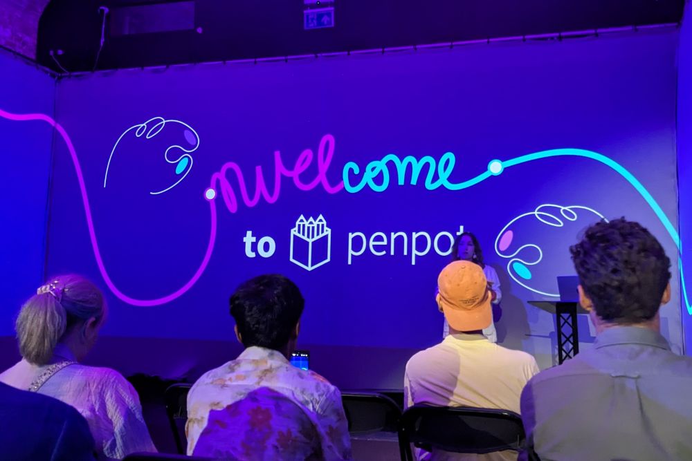 A photo taken at the Penpot social event, I'm sat in the crowd looking towards the screen a few rows ahead of me. The text on the screen reads "Welcome to Penpot" in fun cursive writing.