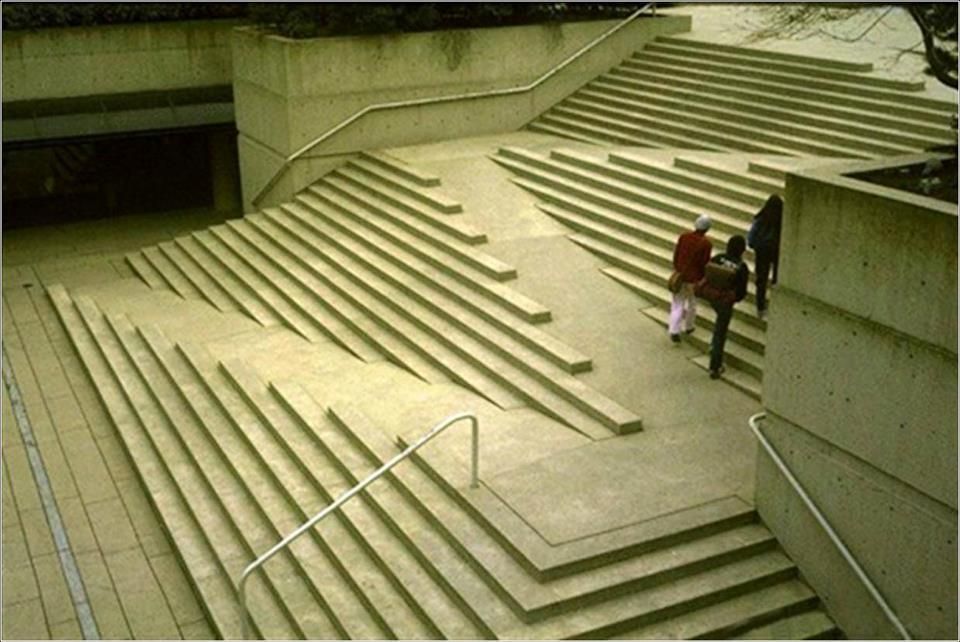 A large concrete staircase with an integrated ramp flanked by shallow, narrow steps that span the width of the structure. Three people are walking up the central ramp, which is embedded within the stair design. The layout is symmetrical but unconventional, blending stairs and ramp in a sculptural, abstract form. Metal handrails are present on the sides.