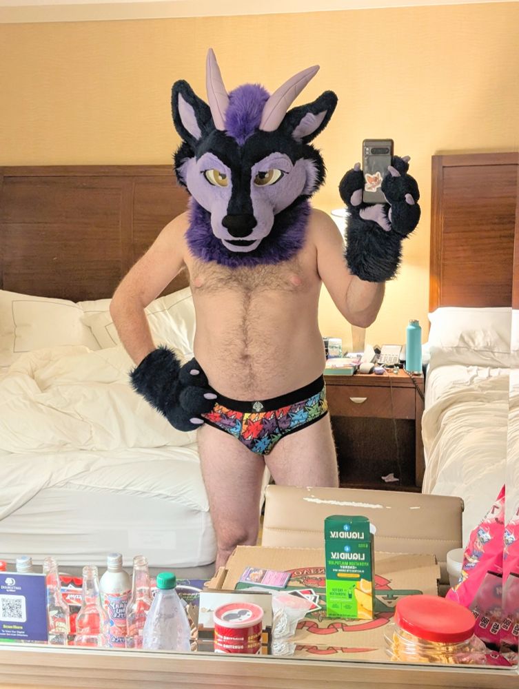 Crow standing in a mirror in fursuit head and undies taking a picture of a very messy hotel desk covered in drinks and take out food