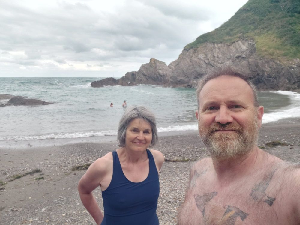 A man and woman in swim gear stand on a beach in a rocky cove. They are smiling for the camera.