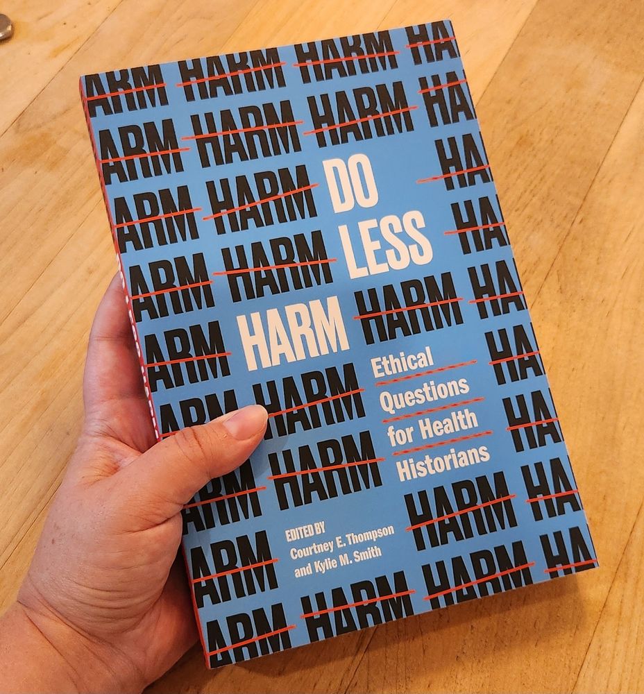 A hand holding a thick paperback book, which has a blue cover with the word "HARM" written in black letters crossed out with a red line repeated as a motif. The title is DO LESS HARM: ETHICAL QUESTIONS FOR HEALTH HISTORIANS edited by Courtney Thompson and Kylie Smith. 