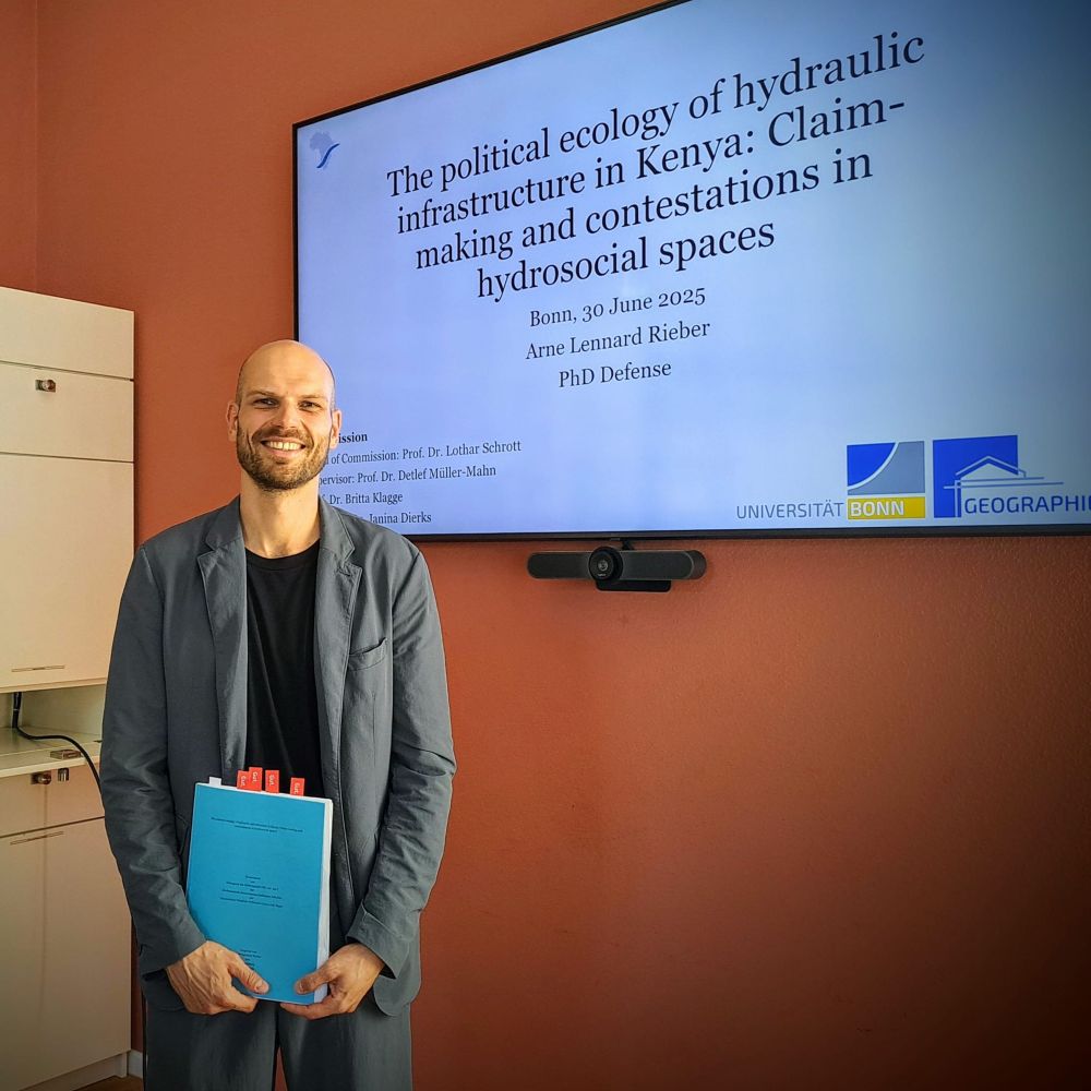 Photo of myself holding my dissertation in front of a screen that shows the title page of my Defense presentation. Title: „The political ecology of hydraulic infrastructure in Kenya: claim-making and contestations in hydrosocial spaces“