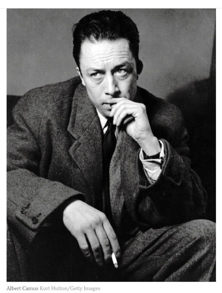 A black and white photo of a pensive looking Albert Camus (1913~1960), dressed in a coat, eyebrows raised, left hand to his lips, and his right hand holding a cigarette between his fingers.
