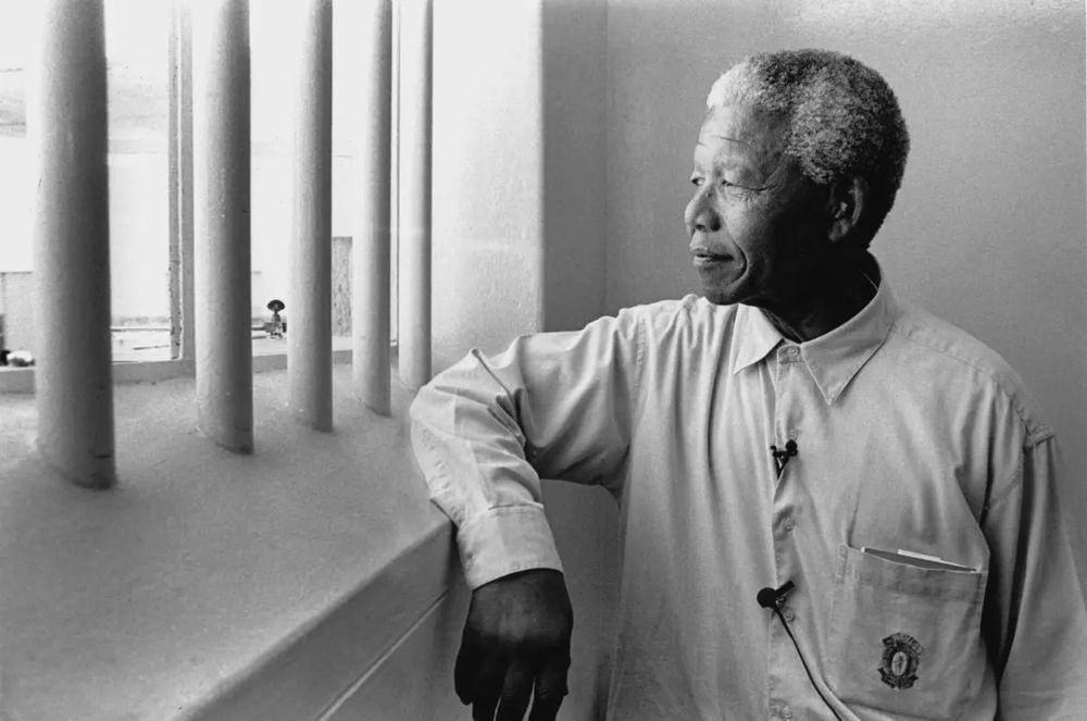 Nelson Mandela, as he looks out through the prison bars, with his right elbow on the ledge.

Then-President Nelson Mandela revisits his South African prison cell on Robben Island, where he spent 18 of his 27 years in prison, in 1994. (Jurgen Schadeberg / Getty Images.