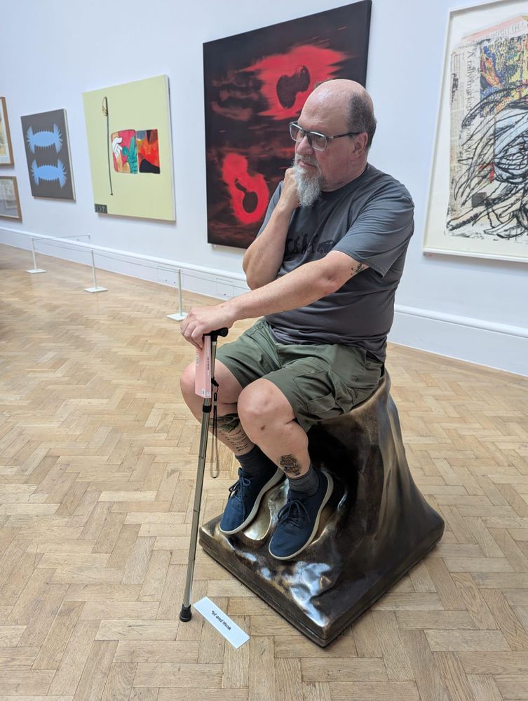 An old man (me) sitting on a bronze sculpture, holding a stick in front of him, in a "Thinking" pose.