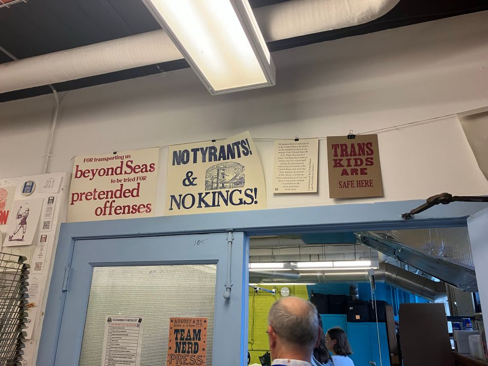 Photo of a wall over an open doorway hung with in letterpress and other kinds of prints, at the Skeuomorph Press. These include: blue ink on cream paper "no tyrants! & no kings!" with illustration of a printing press beribboned with "the people's friend, the tyrant's fire" text; red ink on kraft paper "trans kids are safe here"; and red ink on cream paper "for transporting us beyond seas to be tried for pretended offenses"