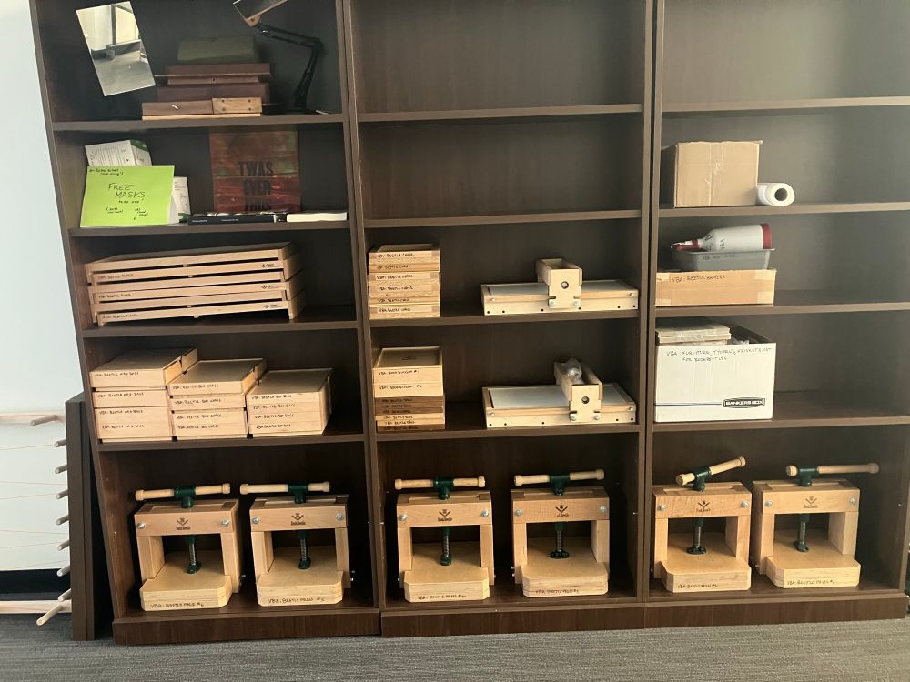 Photo of 3 tall dark-wood bookshelves, bearing 6 BookBeetle tabletop printing presses, a Provisional Press cylinder tabletop press, and multiple boxes of BookBlossom type, all neatly organized