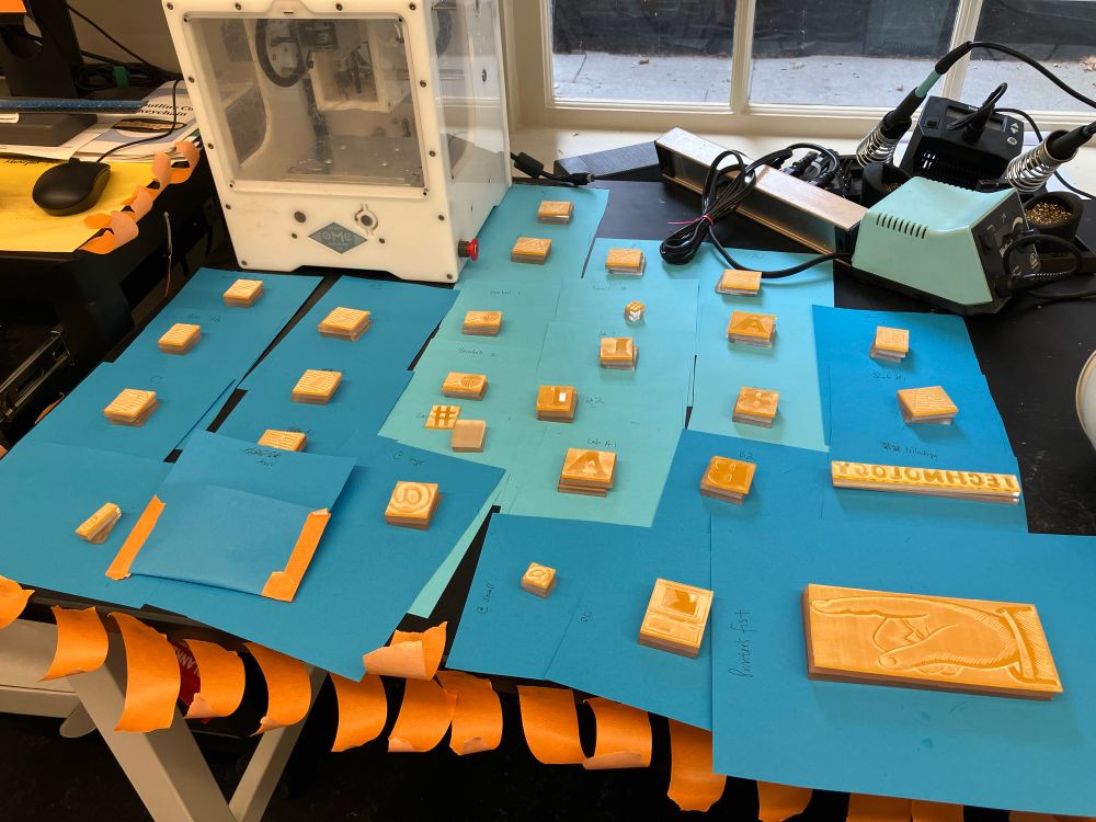 Photo of orange acrylic lasercut sheets stacked on frosted acrylic mounts of the same size, to be glued together to raise them to printing type high. All are reflected backwards so they print the right direction. There's a mix of large Cooper Black letters and images such as a large pointing hand. Each suit on its own piece of blue paper to help keep the mounts of the same size together.