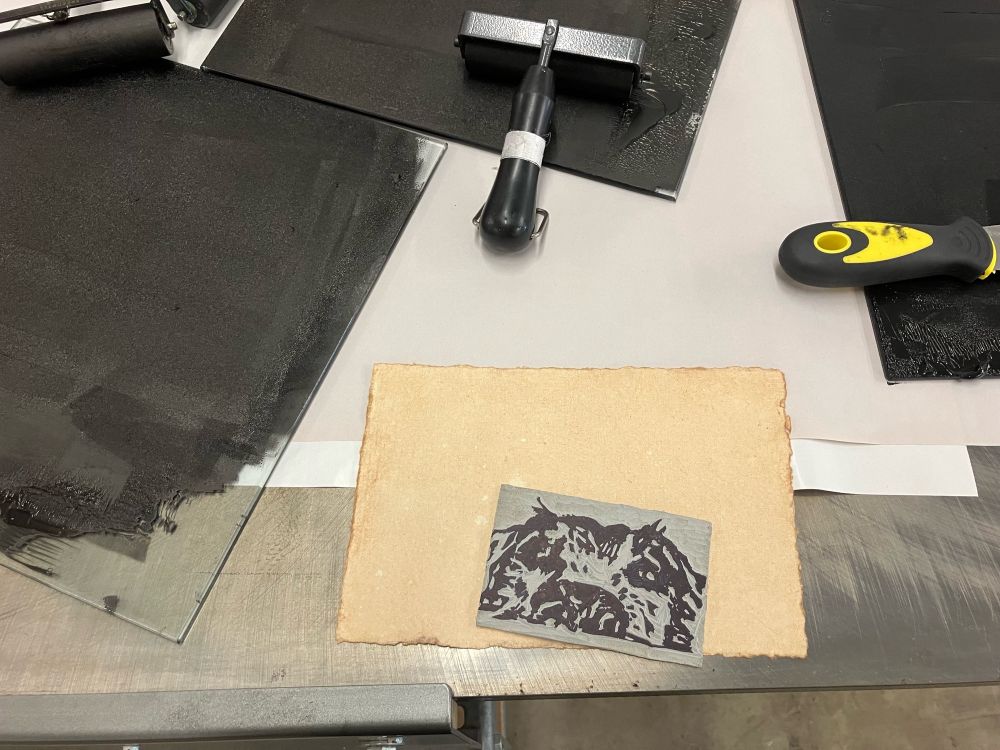 Photo of a linoleum carved block of a dog head based on art by Jes Raymond, rolled in black ink. In the background are glass trays covered in black ink, and printing roller tools (brayers)