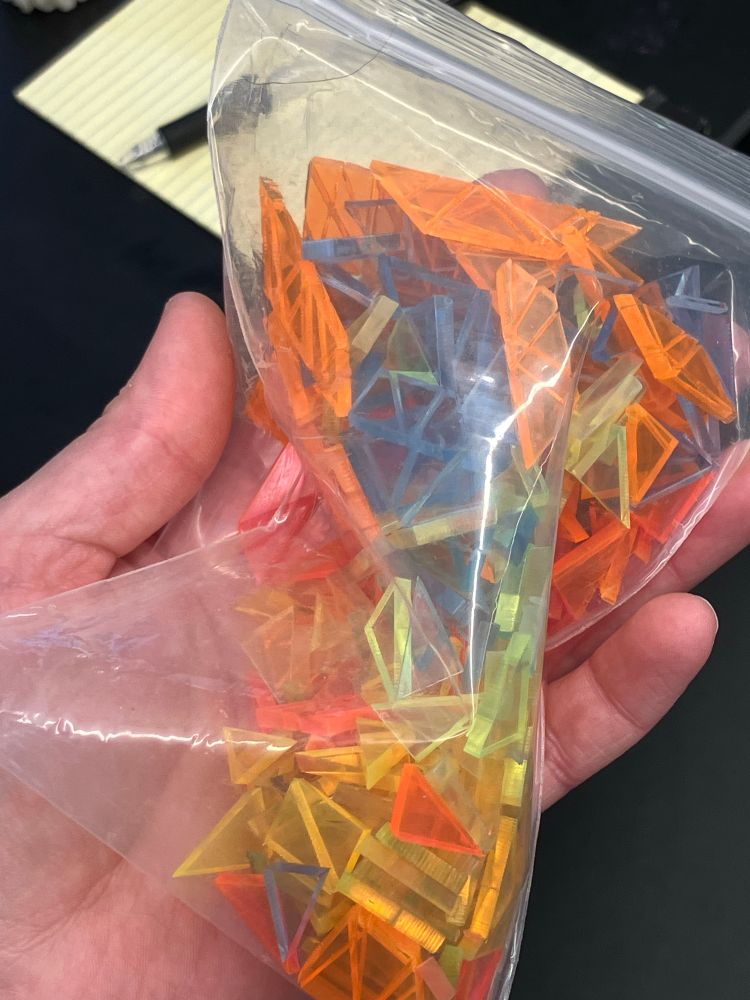 Photo of a hand holding a baggie full of tiny neon shards (acrylic triangles cut with a lasercutter from neon transparent acrylic in various colors)