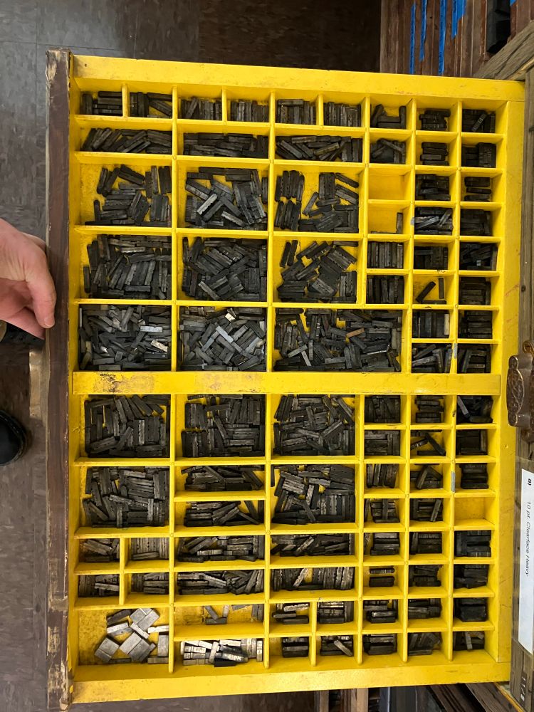 Photo of a letterpress typecase painted bright yellow; it looks like a wide shallow painted-wood drawer with 60+ different small niches for various pieces of type