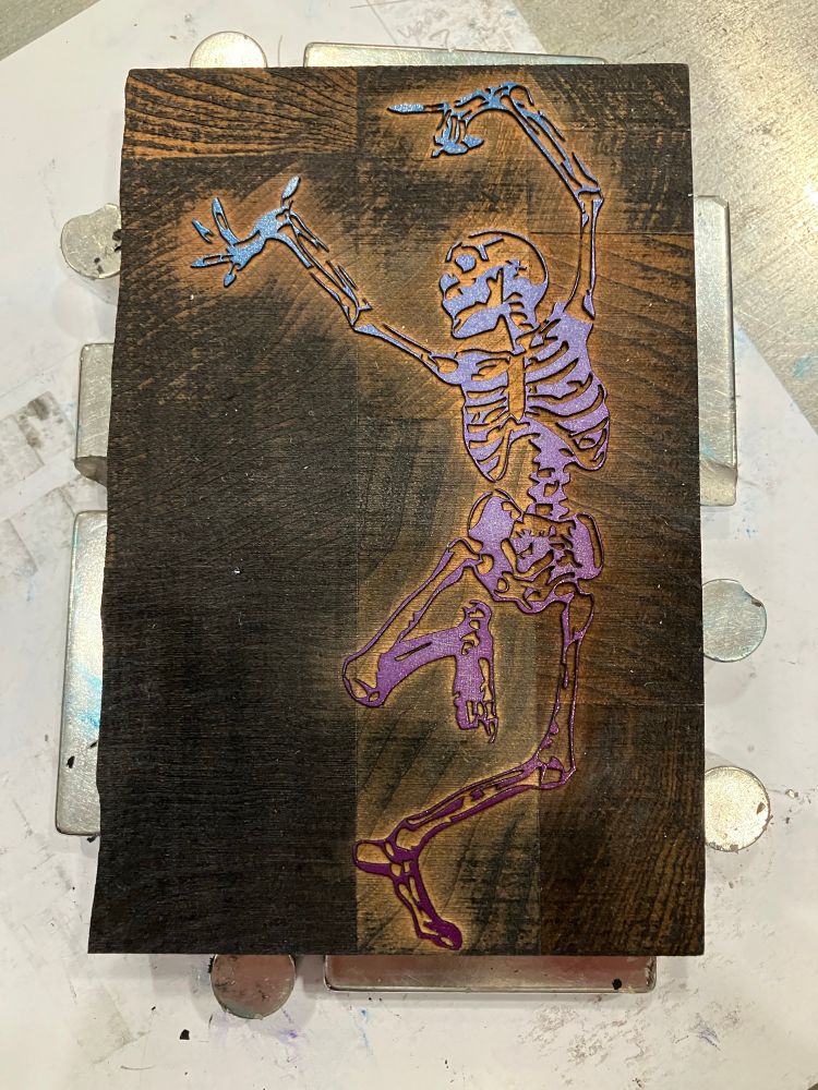 Photo of a wood printing block of a danse macabre skeleton inked in a gradient of red, pink, light blue, blue ink