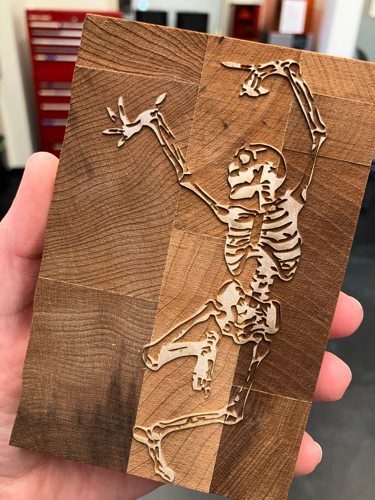 Photo of a wood block cut so an image of a historical "danse macabre" style dancing skeleton cavorts, raised from its surface, for printing