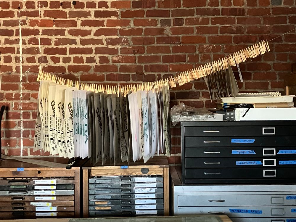 Photo of a drying line full of paper letterpress prints on white and cream parker and brown chipboard, hung using wood clothespins. Some letterpress typecases, flat files, and an exposed red brick wall are in the background.