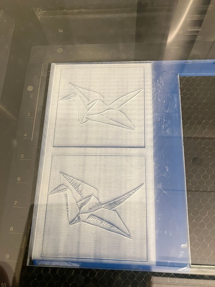 Photo of rectangle of clear acrylic that's in a lasercutter, with two squares cut into it and on each square a drawing of an origami crane etched so it's raised off the surface, for use printing with ink on paper