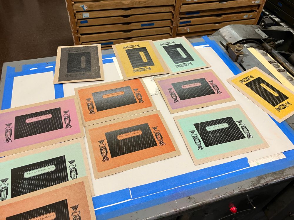 Photo of a table full of letterpress prints made on 9" by 6" brown cardboard stock that's had a slightly gradiented light mauve, robin's egg blue, red, yellow, or black background rectangle inked on it. The foreground is printed in opaque black ink: a diagonally striped rectangle framing a tiny window displaying the tiny text "Rigor's bullshit & you know it"; carytids flank the sides of the rectangle. 