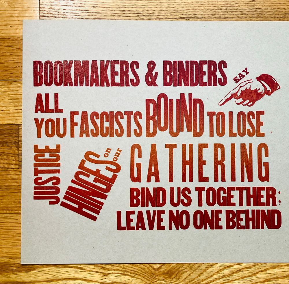 Photo of a letterpress print made with wood and lead type on light brown chipboard, and a gradient of red yellow orange ink for the text. It says "Bookmakers & binders say all you fascists bound to lose / Justice hinges on us gathering / bind us together / leave no one behind". The word "hinges" is set diagonally off the word "justice" to look like a book hinging open
