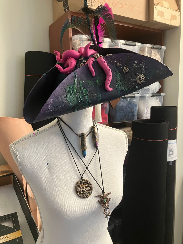 Photo from the Scholars' Lab makerspace of user-made necklaces that look like a bronze medallion, two bronze gem pendant holders, and an elaborate vial holder necklace; and a purple decorated tricorner felt hat with feathers and pink tentacles peeking out from behind the brim