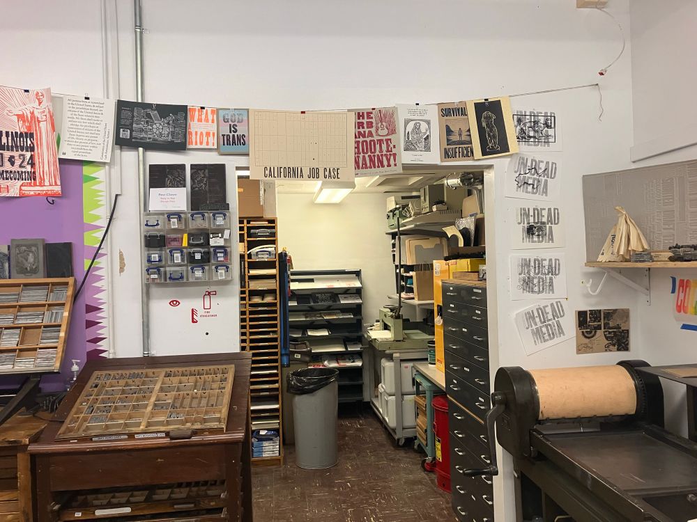 Photo of a wall around an open doorway, covered in letterpress and other kinds of prints, at the Skeuomorph Press. These include: briwn ink on cream paper "California job case" with an illustration of the same, black ink on trans flag pink/white/blue paper "God is trans", and black ink on white paper "undead media" with a large superimposed print of an eye