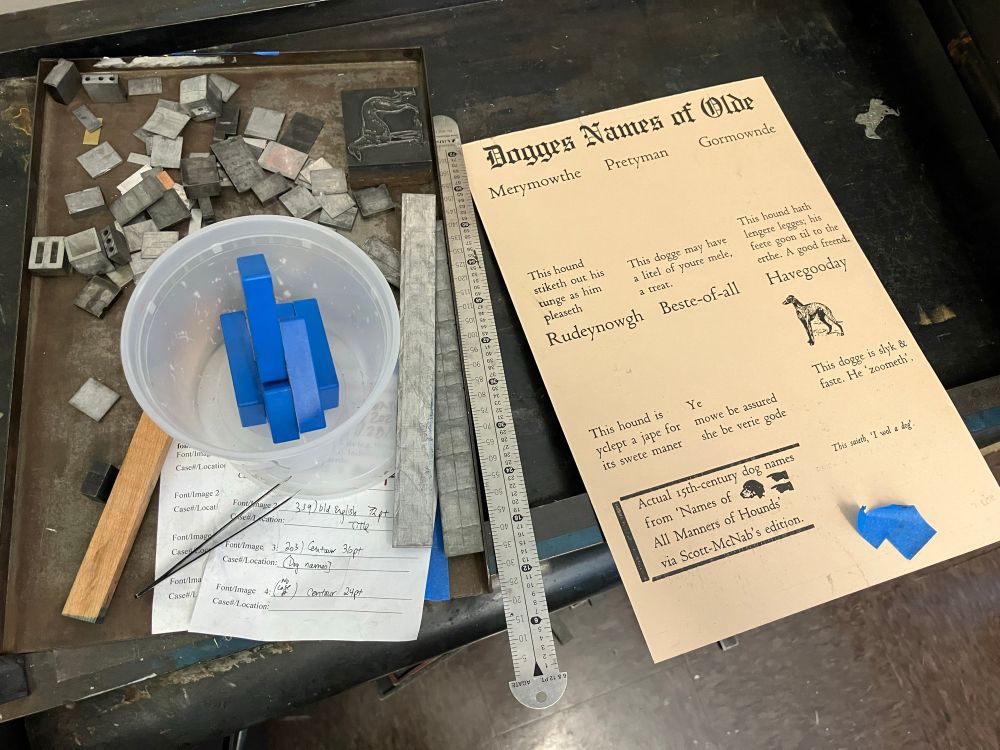 Photo of a half-done version of my letterpress print, next to a metal galley tray containing various letteroress spacing, magnets, and blocks. The print used Old English and Centaur fonts, and various metal and wood dog illustration blocks, inked in black. The text reads in somewhat inaccurate mock late Middle English: "Dogges Names of Olde" followed by names over each of 6 spaces for eventual dog illustrations (Rudynowgh, Gourmande, Merymowthe, Pretyman, Havegooday, Beste-of-all) and captions under each: "This dogge may have a litel of youre mele, a treat." "This hound hath lengere legges; his feete goon til to the erthe. A good freend." "This hound stiketh out his tunge as him pleaseth." “This hound is yclept a jape for its swete maner.” “Ye mowe be assured she be verie gode.” “This dogge is slyk & faste. He ‘zoometh’.” At the bottom is a bordered section that says “Actual 15th-century dog anmes from ‘Names of All Manners of Hounds’ via Scott-McNab’s edition.” To the side of this, smaller text in italics says “This saieth, ‘I wol a dog.’” 