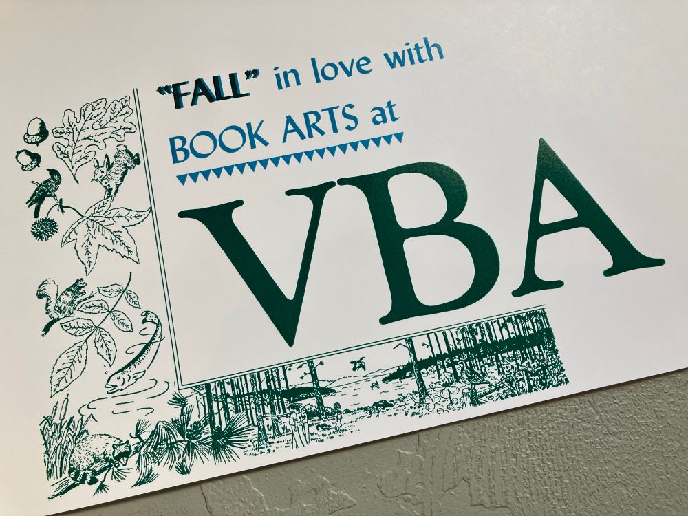Photo of a letterpress print in teal and green inks on cream paper. It says "'Fall' in love with book arts at VBA", inside an L-shaped border showing a detailed autumnal forest lake scene