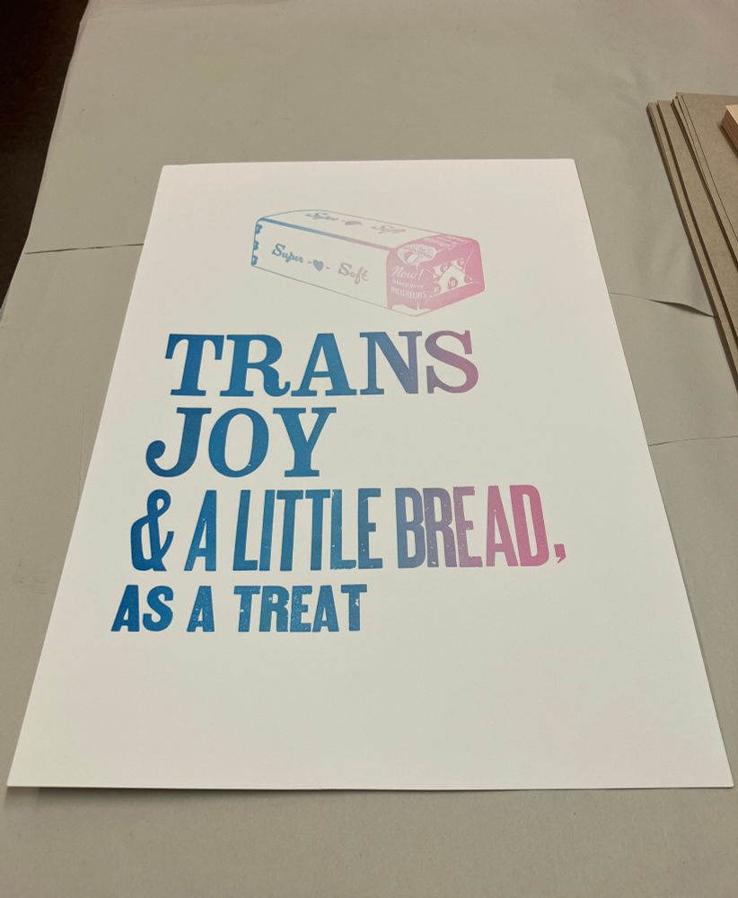 Photo of a letterpress print using caps serif wood type that says "Trans Joy", followed by smaller sans serif text that says "& a little bread, as a treat"; all under a large sandwich bread illustration print; inked in a trans-flag blue to pink gradient. 