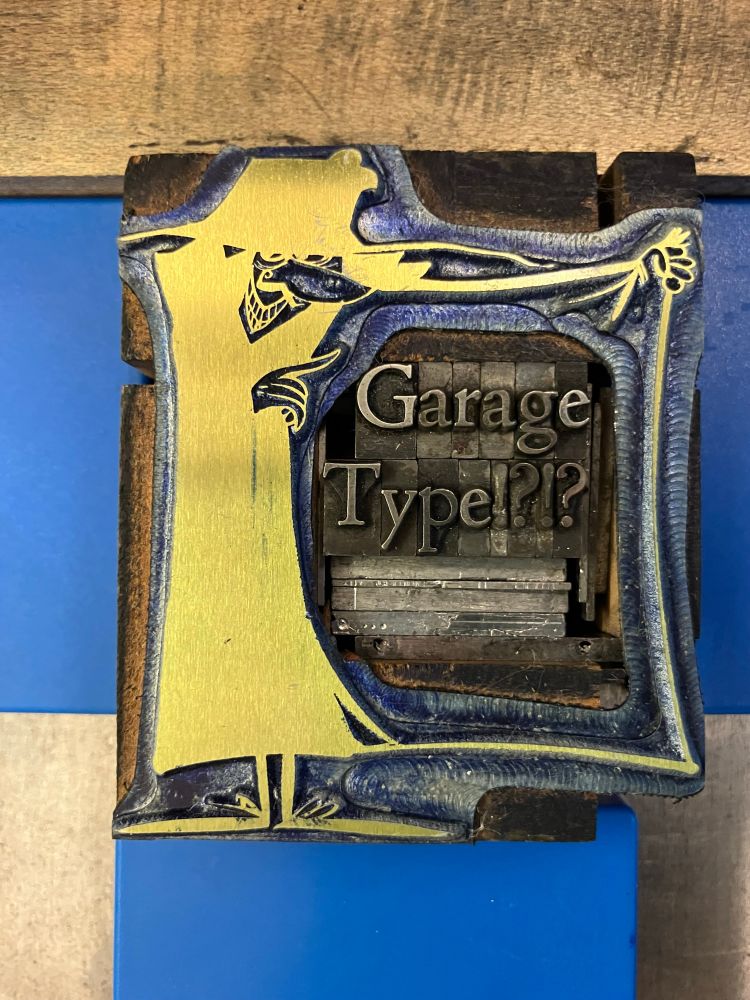Photo of a mortised (carved-out space to insert type) letterpress block showing a cartoon of a shifty person trying to sell you something from inside one side of an opened trechcoat; inside the trenchcoat is arranged the type "Garage type!?!?" in a serif font. 