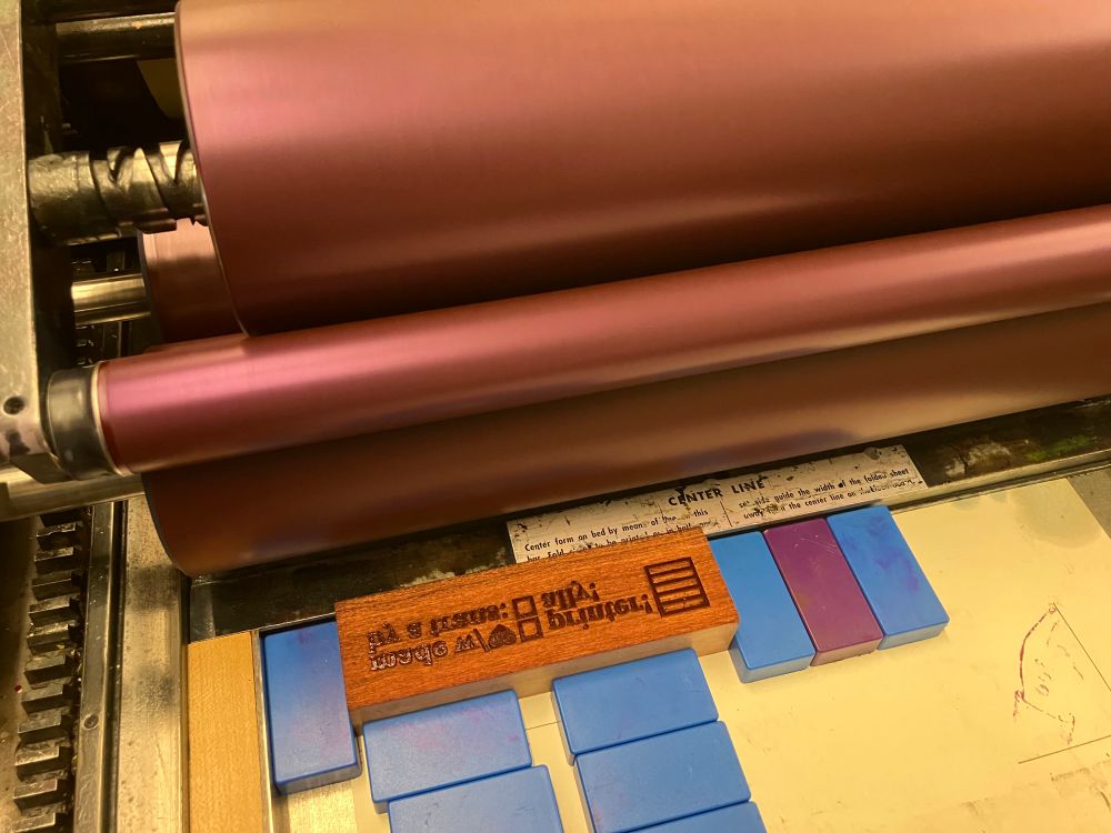 Photo of a letterpress printer with rollers inked in a deep burgundy bullet, and in the press bed amid blue magnets, a cherry wood block lasercut to say "made w/💜 by a trans (checkbox) printer (checkbox) ally" and the outline of a trans flag
