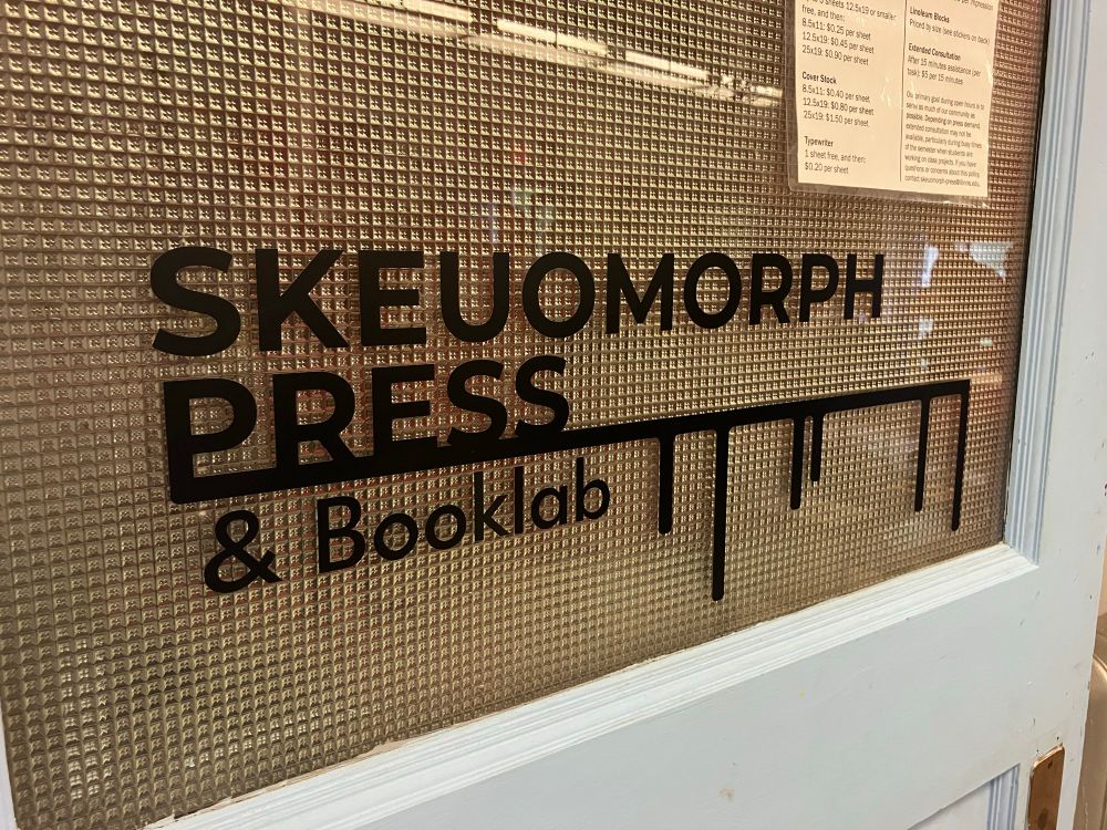 Photo of a white-painted wooden door with a frosted grid window in its upper half, which has a black adhesive sign on the glass that says "Skeuomorph Press & BookLab"; the is an underline under the first two words that has some decorative drip details on it