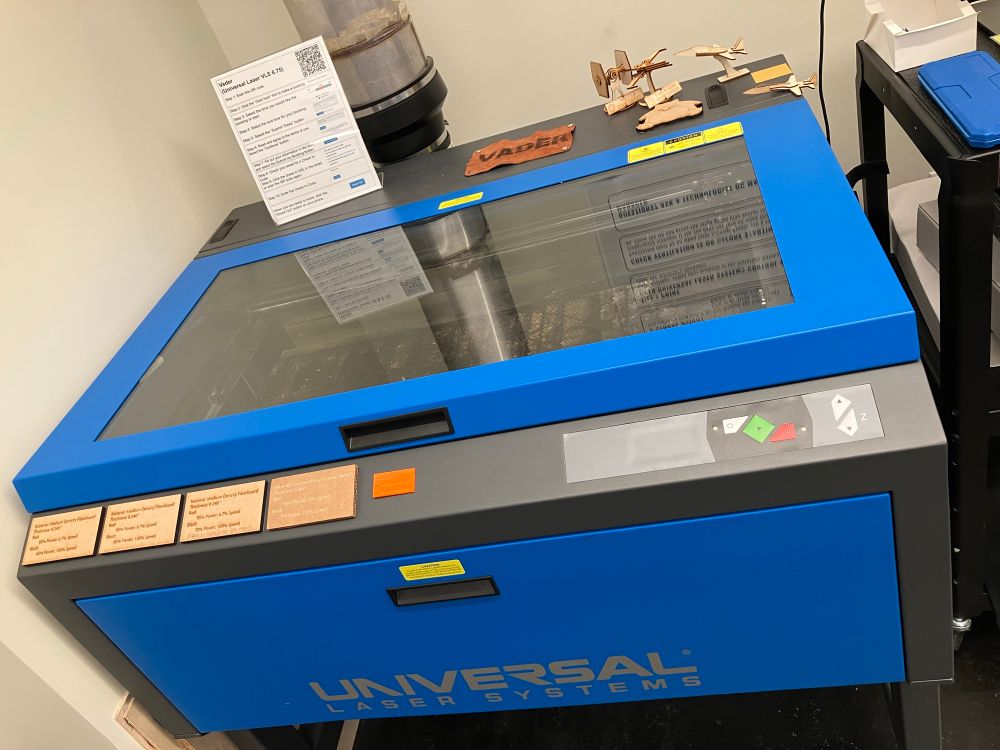 Photo of the Scholars' Lab Makerspace lasercutter, named Vader, a large oven-sized royal blue container with a pipe to ventilate it and glass top to see the laser working through. Some lasercut wood airplanes and Star Wars ships, and a laser engraved piece of leather that dates "Vader", are visible on the back of the machine