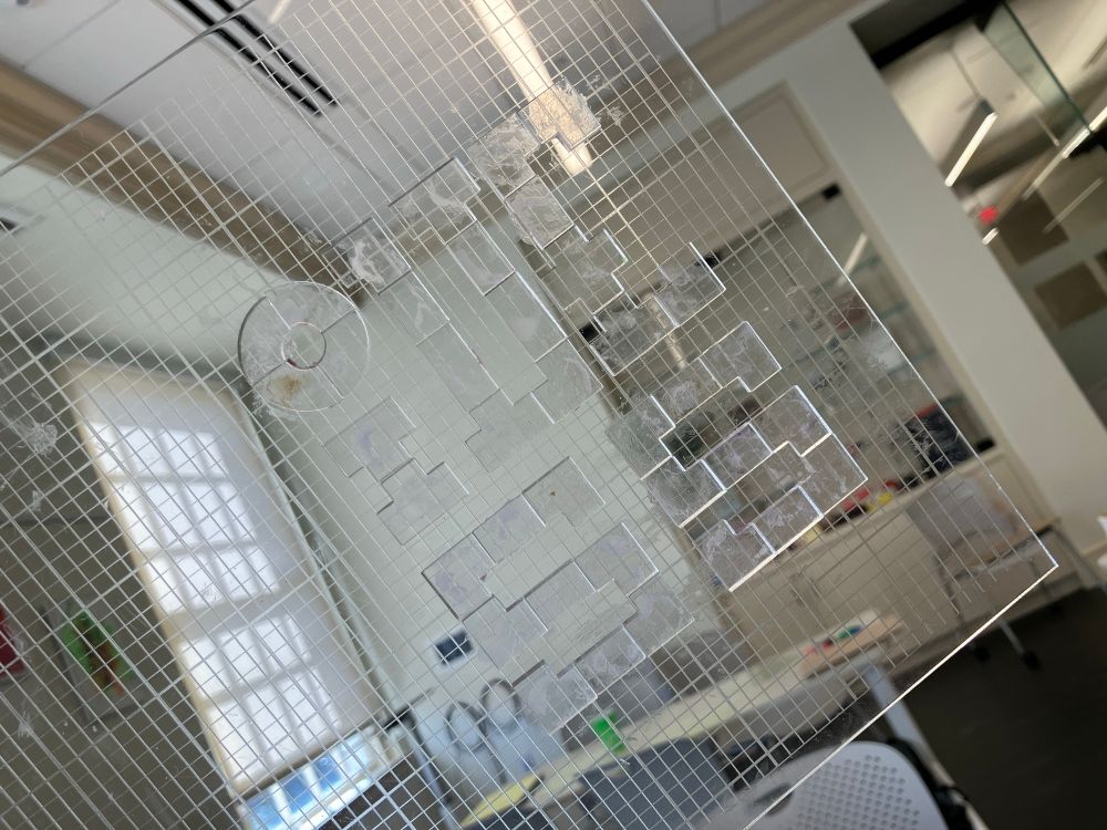 Photo of a printing block by Josef Beery; it's clear acrylic small shapes, arranged into a an artistic layout of the words "zine zone" (backwards so it prints readably), that can be inked and printed on paper. Held up to show the light coming through. 