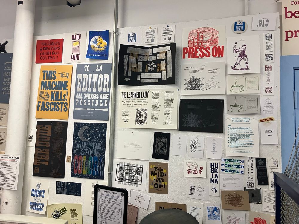 Photo of a wall covered in letterpress and other kinds of prints, at the Skeuomorph Press. These include: navy ink on maize paper "this machine kills fascists", white/silver ink on blue paper "to an editor all things are possible", and rainbow ink on midnight blue "when I dream, I dream in color"