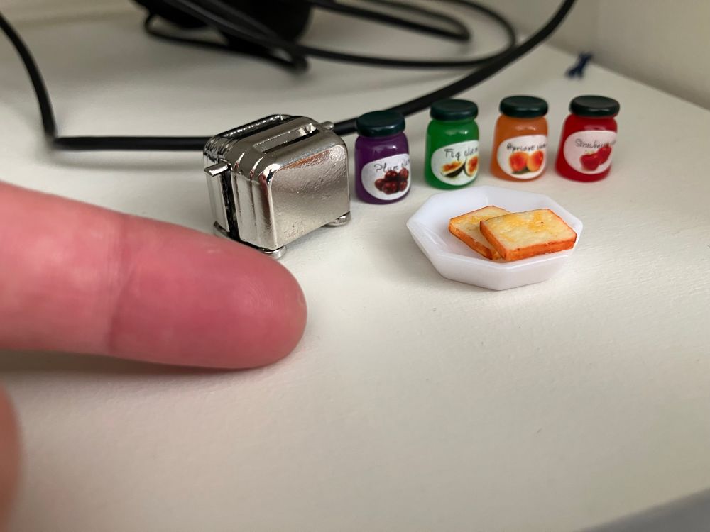 Close up photo of tiny doll house items on a white shelf: 4 tiny jam jars with fruit labels and colors, a chrome toaster, and a plate with two buttered pieces of toast. My index finger is shown next to the items to demonstrate their dollsized scale. 