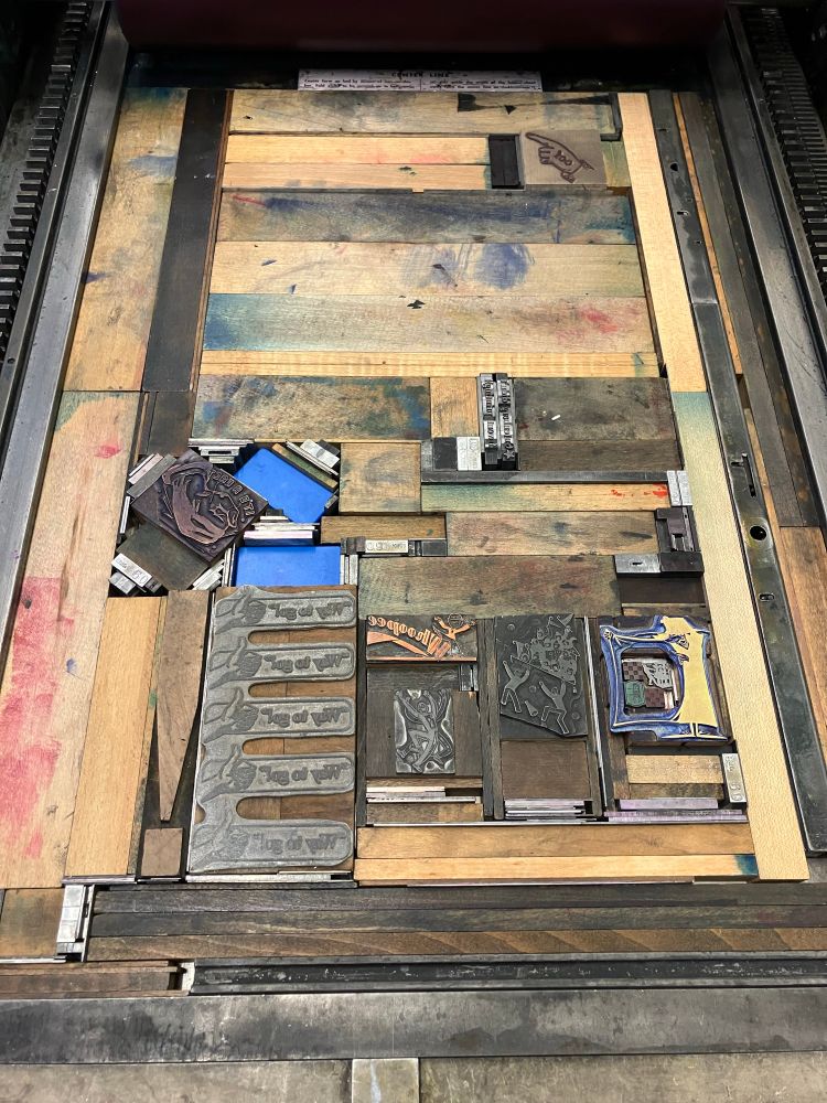 Photo of a letterpress print bed packed with wood and metal blocks to form a solid rectangle around a variety of metal illustration blocks such as a shady man holding open his trenchcoat to show he is selling old print presses, and a 5-times-repeated motif of a hand giving a thumbs up next to the text "way to go!"