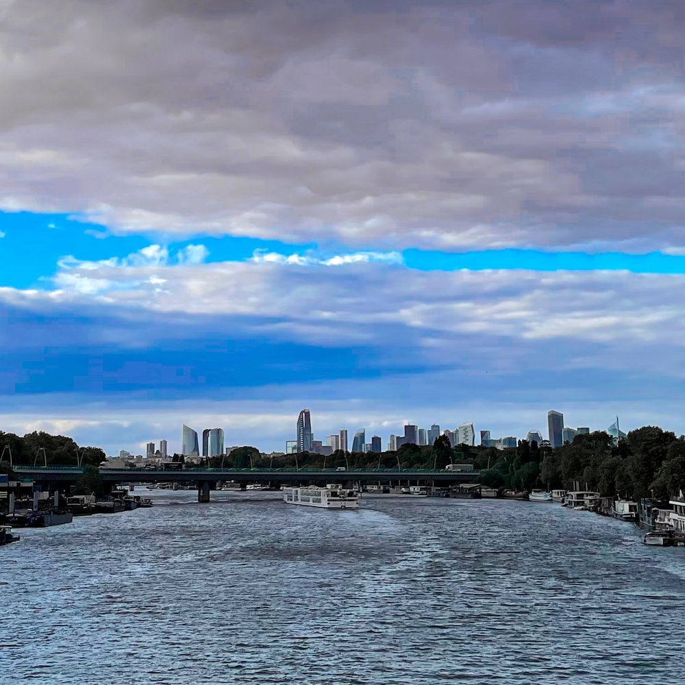 Sous le ciel nuageux mais néanmoins doté d’une bande bleue du meilleur goût, la Seine où navigue un bateau battu par les flots et au loin les tours du quartier de La Défense 
