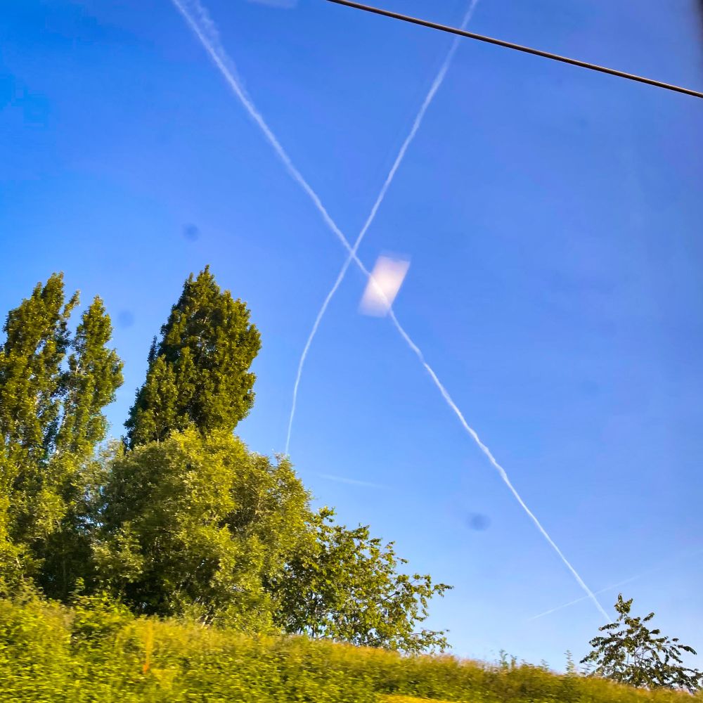 Le ciel vu du TGV, avec des reflets pourris dans la vitre sale, deux chemtrails forment un X suspect dans ledit ciel, à noter des arbres et arbustes au premier plan, ainsi qu’il câble électrique