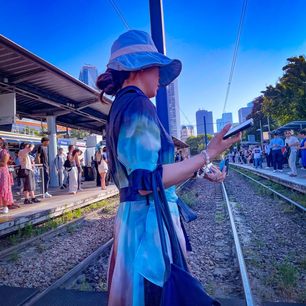 Une dame en robe bleu et chapeau assorti traverse les voies du tramway dont les quais sont bondés, les tours de La Défense en arrière-plan 