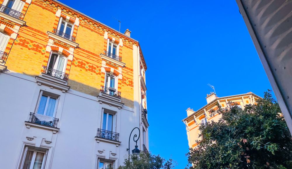 Le ciel est très bleu au-dessus de la banlieue, le soleil levant éclaire d’une lumière orangée le haut des immeubles en briques claires, des arbres et un réverbère complètent élégamment le paysage urbain, ainsi qu’un bout de volet antédiluvien sur la droite de la photo 
