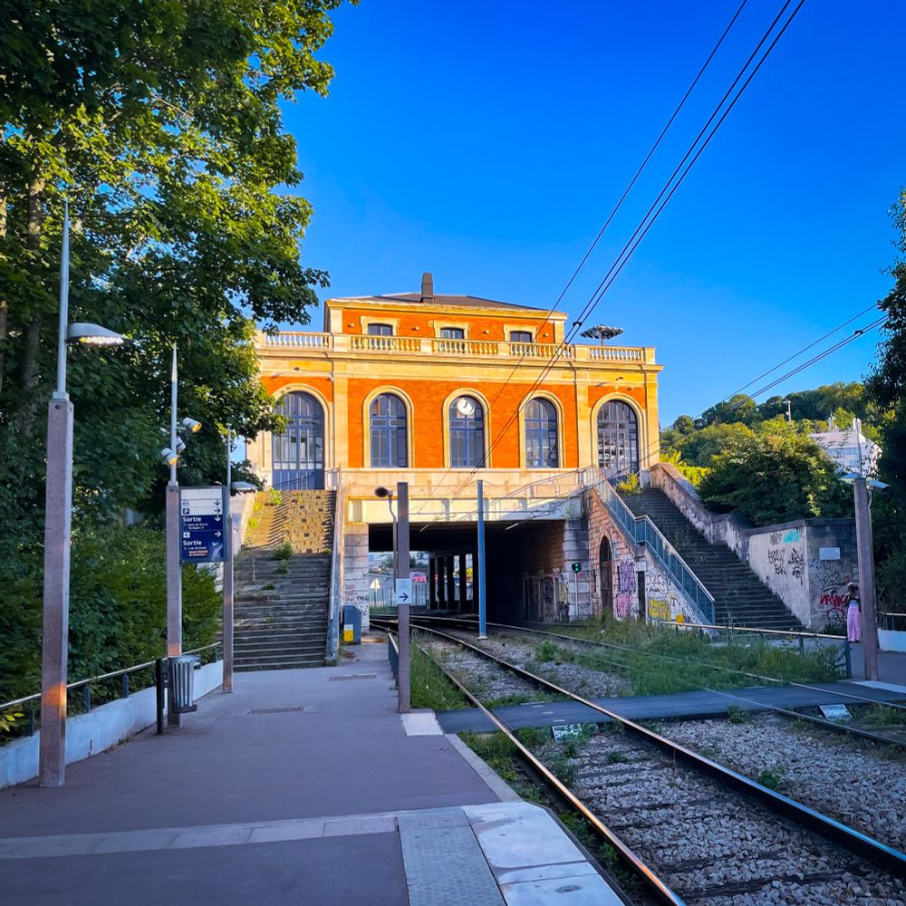 Station du tramway à Sèvres ; le vieux bâtiment de la gare qui enjambe les voies dudit tramway est baigné par la lumière orangée du soir, le ciel est bleu