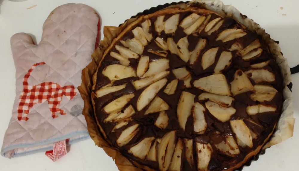 A photo of a chocolate tart with pear sliced arranged in circles on it, with a light-blue kitchen gloves on the side that has a cat on it made of red and white squares.